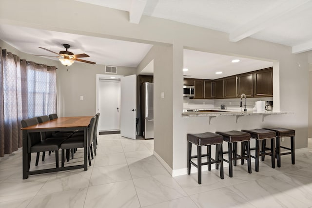 kitchen with light stone counters, beamed ceiling, kitchen peninsula, dark brown cabinets, and appliances with stainless steel finishes