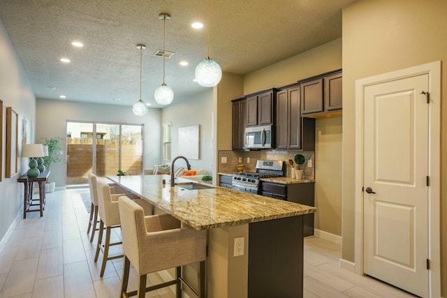 kitchen with sink, hanging light fixtures, stainless steel appliances, light stone counters, and a center island with sink