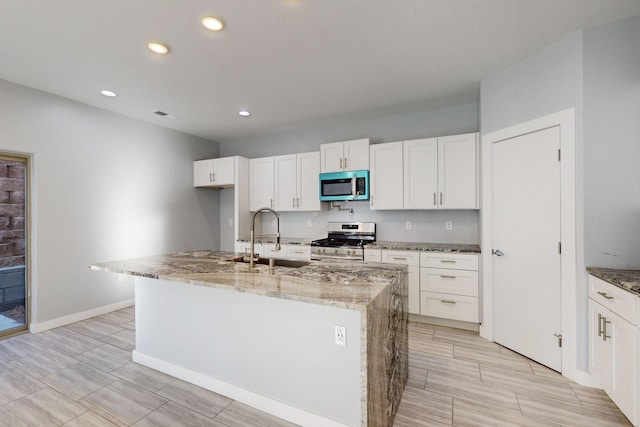 kitchen with stainless steel appliances, an island with sink, light stone countertops, and white cabinetry