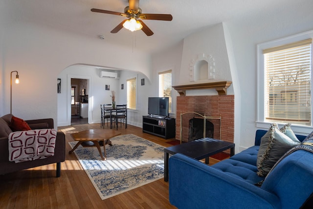 living room with a fireplace, wood-type flooring, a wall unit AC, and ceiling fan