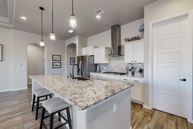 kitchen featuring white cabinetry, wall chimney range hood, decorative light fixtures, a kitchen island with sink, and appliances with stainless steel finishes