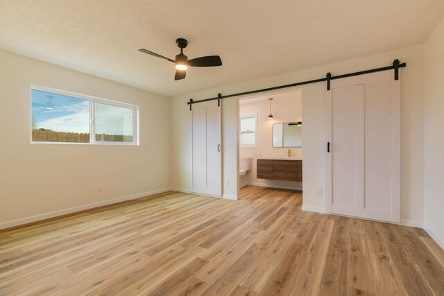 unfurnished bedroom with ensuite bath, a barn door, ceiling fan, and light wood-type flooring