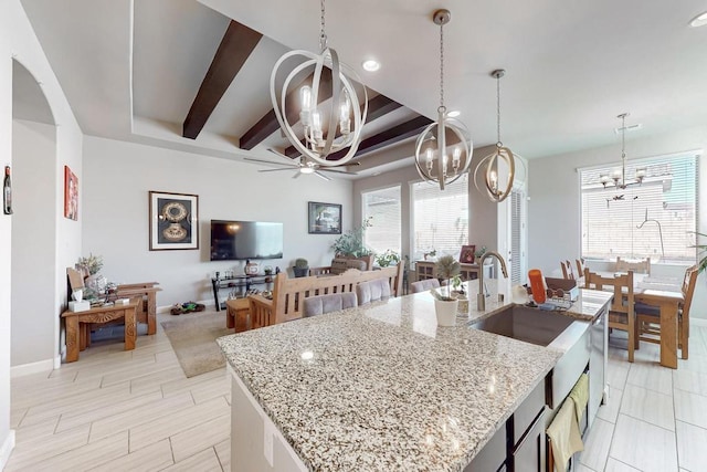 kitchen featuring hanging light fixtures, light stone counters, beamed ceiling, a center island with sink, and ceiling fan with notable chandelier