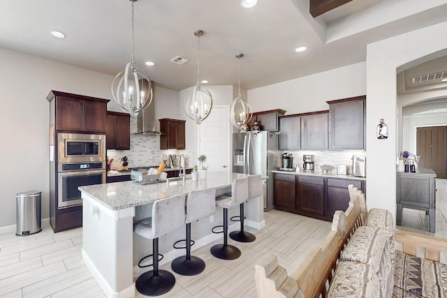 kitchen with appliances with stainless steel finishes, backsplash, light stone counters, decorative light fixtures, and a center island with sink