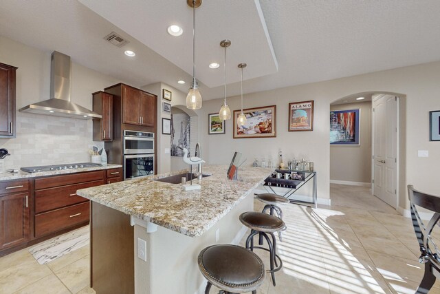 kitchen with sink, appliances with stainless steel finishes, an island with sink, a kitchen bar, and wall chimney exhaust hood