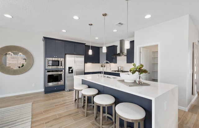 kitchen featuring pendant lighting, stainless steel appliances, a center island with sink, and wall chimney range hood