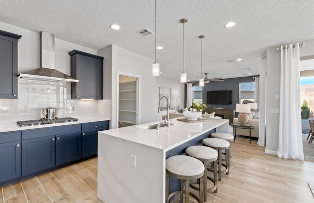 kitchen featuring pendant lighting, a center island with sink, wall chimney range hood, sink, and stainless steel gas cooktop