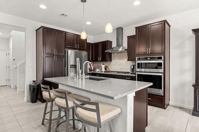 kitchen with a center island with sink, a kitchen breakfast bar, wall chimney range hood, sink, and appliances with stainless steel finishes