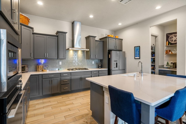 kitchen featuring stainless steel appliances, an island with sink, a kitchen bar, sink, and wall chimney range hood