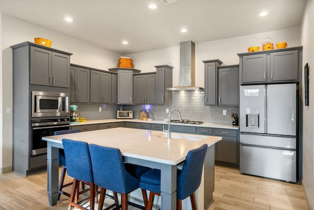 kitchen featuring wall chimney exhaust hood, a center island with sink, a kitchen bar, gray cabinetry, and appliances with stainless steel finishes