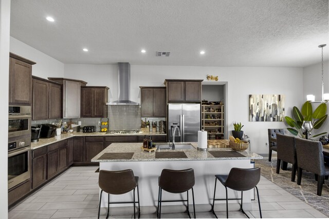 kitchen with stainless steel appliances, wall chimney exhaust hood, a kitchen breakfast bar, decorative backsplash, and a center island with sink