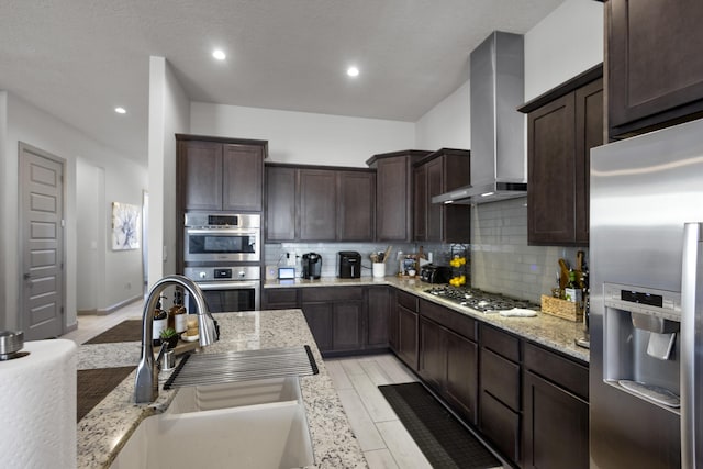 kitchen featuring a sink, dark brown cabinets, appliances with stainless steel finishes, wall chimney exhaust hood, and tasteful backsplash