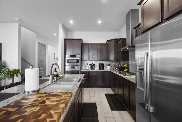 kitchen featuring stainless steel appliances, a sink, dark brown cabinets, wall chimney range hood, and backsplash