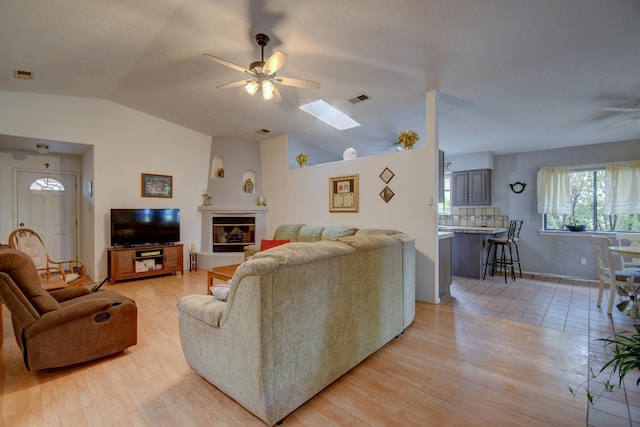 living room featuring ceiling fan, a fireplace, lofted ceiling with skylight, and light wood-type flooring