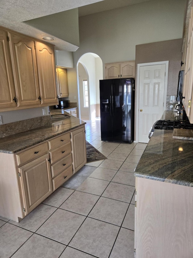 kitchen with light tile patterned floors, dark stone countertops, a high ceiling, black fridge with ice dispenser, and light brown cabinets