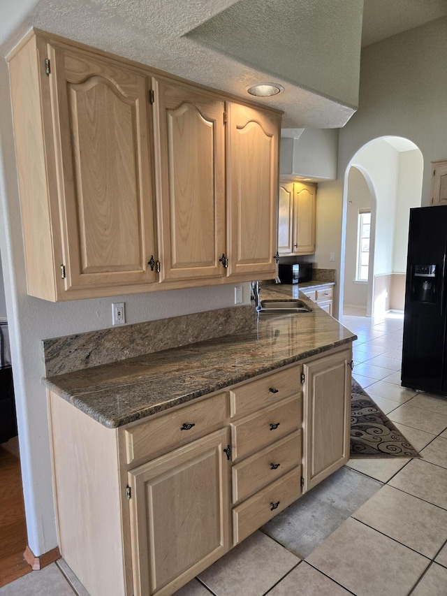 kitchen with light tile patterned floors, light brown cabinetry, black refrigerator with ice dispenser, and dark stone countertops