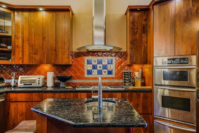 kitchen with an island with sink, stainless steel appliances, dark stone counters, and wall chimney range hood