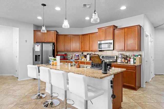 kitchen with appliances with stainless steel finishes, a kitchen bar, an island with sink, hanging light fixtures, and light stone counters