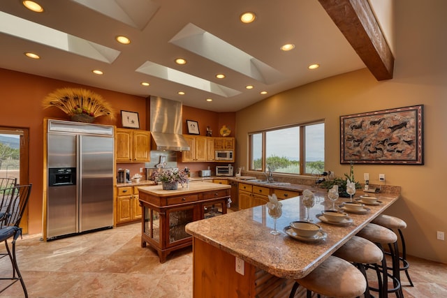 kitchen featuring a skylight, built in appliances, kitchen peninsula, beam ceiling, and wall chimney range hood