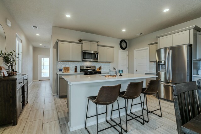 kitchen featuring a breakfast bar area, backsplash, a kitchen island with sink, gray cabinetry, and appliances with stainless steel finishes