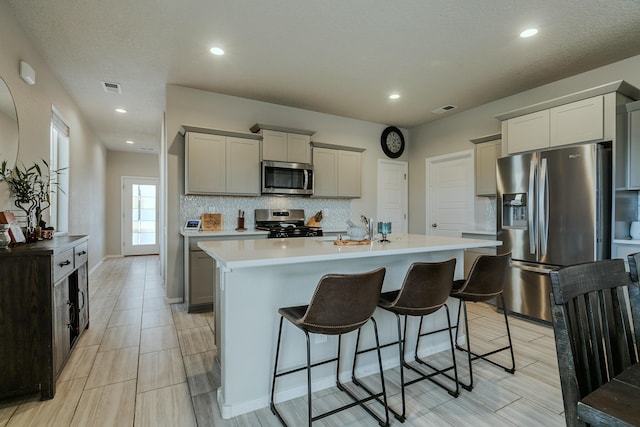 kitchen featuring appliances with stainless steel finishes, backsplash, gray cabinets, a breakfast bar, and a center island with sink