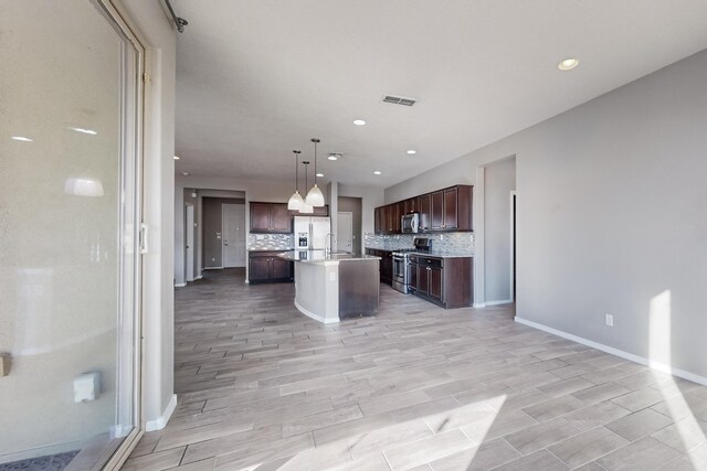 kitchen with an island with sink, appliances with stainless steel finishes, tasteful backsplash, decorative light fixtures, and dark brown cabinetry