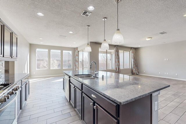 kitchen featuring decorative light fixtures, a center island with sink, dark brown cabinets, sink, and dark stone counters