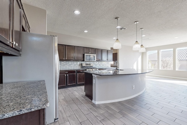 kitchen featuring hanging light fixtures, dark stone counters, stainless steel appliances, and a center island with sink