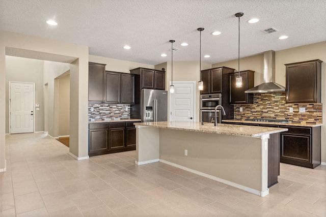 kitchen with backsplash, wall chimney range hood, hanging light fixtures, appliances with stainless steel finishes, and an island with sink