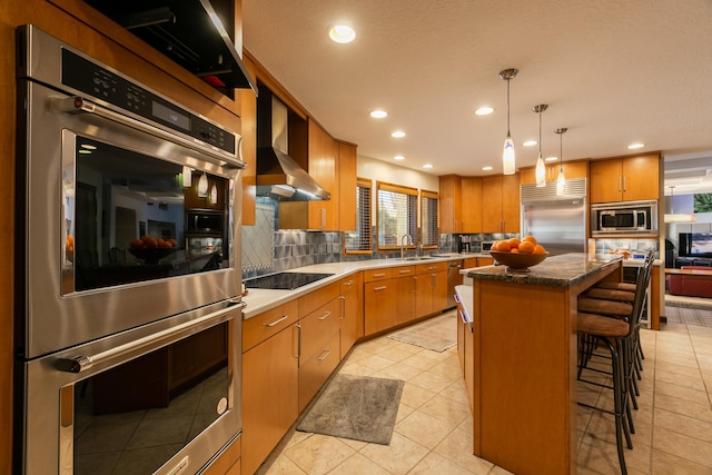 kitchen with a kitchen island, pendant lighting, a breakfast bar area, built in appliances, and wall chimney exhaust hood