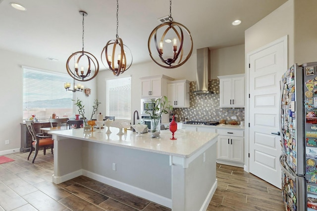 kitchen with appliances with stainless steel finishes, decorative light fixtures, wall chimney range hood, white cabinetry, and a center island with sink