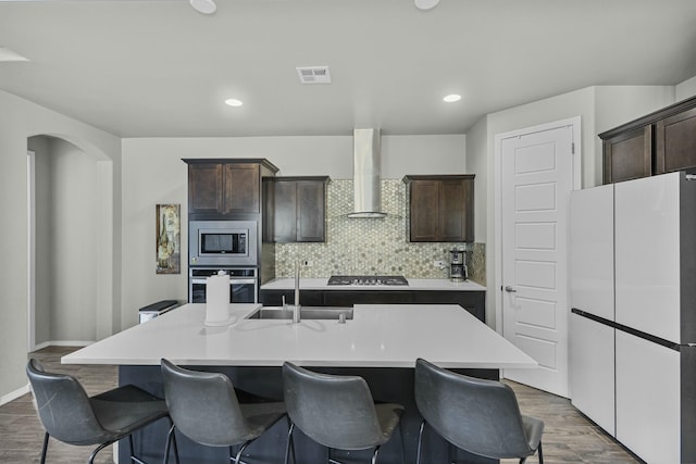 kitchen featuring wall chimney range hood, stainless steel appliances, tasteful backsplash, a kitchen island with sink, and dark brown cabinets