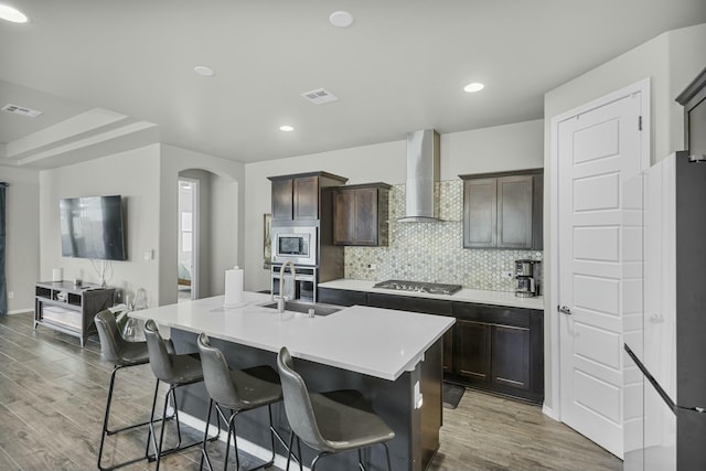 kitchen with stainless steel appliances, a kitchen island with sink, wood-type flooring, wall chimney exhaust hood, and dark brown cabinetry