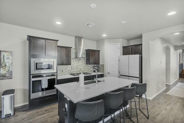 kitchen featuring wall chimney range hood, appliances with stainless steel finishes, dark brown cabinets, and an island with sink