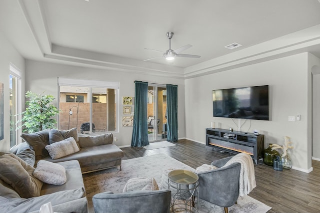 living room featuring visible vents, a glass covered fireplace, wood finished floors, baseboards, and a raised ceiling