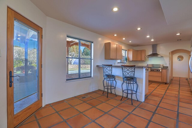 kitchen featuring kitchen peninsula, a breakfast bar area, tile patterned flooring, light brown cabinetry, and wall chimney range hood