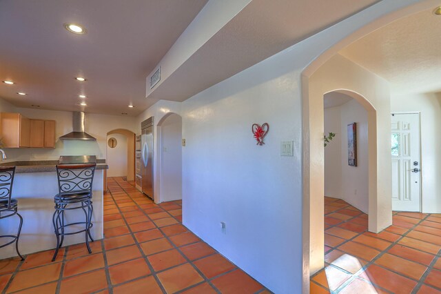 kitchen with built in fridge, wall chimney exhaust hood, light brown cabinets, tile patterned floors, and a kitchen breakfast bar