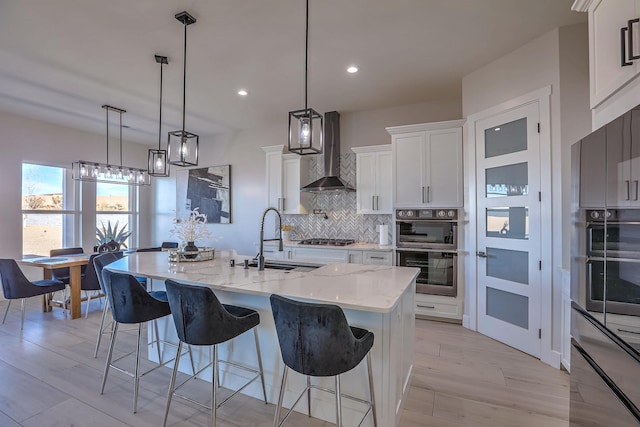 kitchen with decorative light fixtures, a large island with sink, sink, white cabinetry, and wall chimney exhaust hood