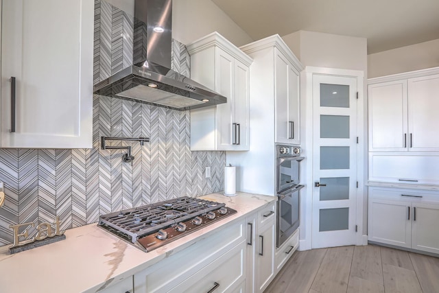 kitchen featuring light stone countertops, white cabinets, appliances with stainless steel finishes, wall chimney range hood, and decorative backsplash