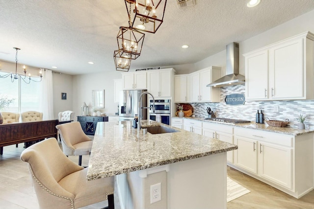 kitchen with stainless steel appliances, wall chimney exhaust hood, a center island with sink, and hanging light fixtures