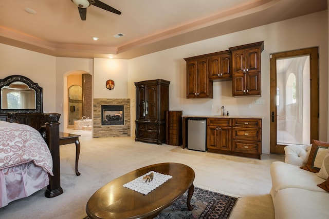 carpeted bedroom featuring ceiling fan, a tray ceiling, ensuite bath, a fireplace, and refrigerator