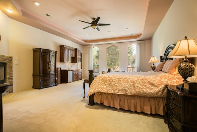 bedroom featuring ceiling fan, light colored carpet, a raised ceiling, and a stone fireplace