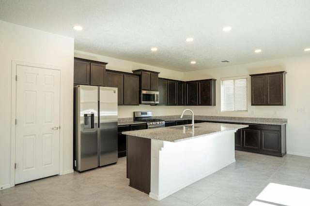 kitchen with sink, stainless steel appliances, light stone counters, a center island with sink, and dark brown cabinetry