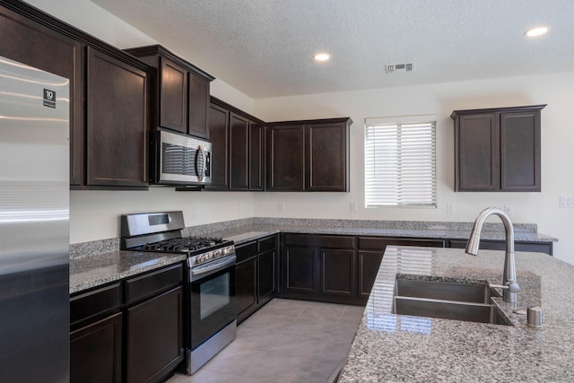 kitchen with a textured ceiling, appliances with stainless steel finishes, sink, light stone counters, and dark brown cabinets