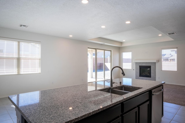 kitchen with light stone countertops, dishwasher, sink, a center island with sink, and a tray ceiling