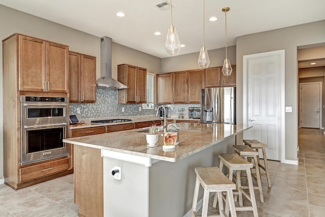 kitchen with sink, stainless steel appliances, light stone counters, an island with sink, and wall chimney exhaust hood