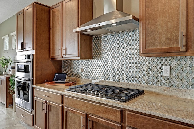 kitchen with wall chimney range hood, light tile patterned floors, stainless steel appliances, light stone counters, and decorative backsplash