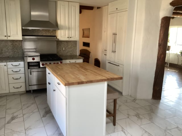 kitchen with wall chimney exhaust hood, white cabinetry, wooden counters, stainless steel stove, and decorative backsplash