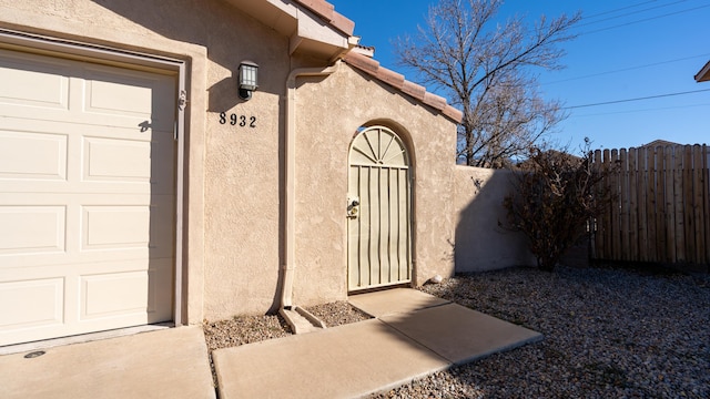 view of exterior entry featuring a garage, a tiled roof, fence, and stucco siding