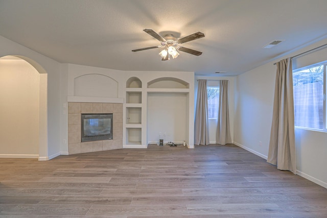 unfurnished living room featuring a tile fireplace, ceiling fan, wood-type flooring, a textured ceiling, and built in shelves